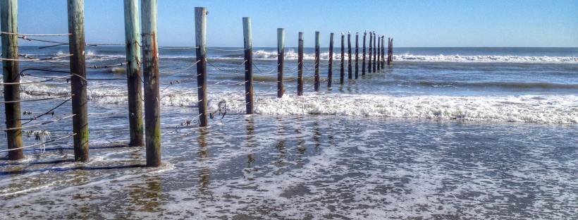A wave crashing into a beach at Outerbank in North Carolina. There's a fence going into the water, presumably keeping people out.