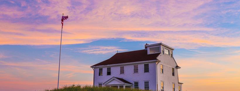 A white two-storey house on the top of a hill in Cape Cod with an American flag blowing in the wind at sunset