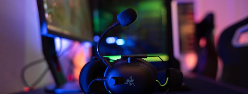 A pair of gamer headphones sit on a desk with two computer monitors in the background, all in dimly lit room with neon lights.