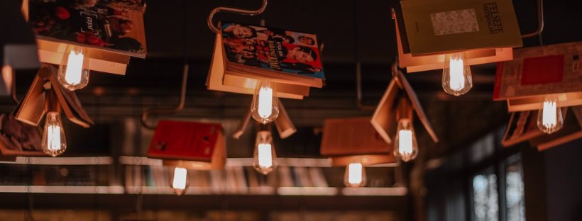 Edison bulbs are hanging from the ceiling in a bookstore. The bulbs have books draped over them so they kind of look like butterflies or little houses.