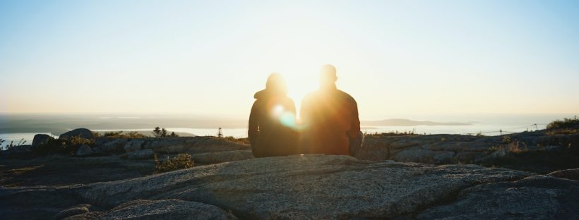 A photo of a couple sitting at the top of a rocky hill at sunrise. They have their backs to us and the sun is right between them, so we can barely make them out.