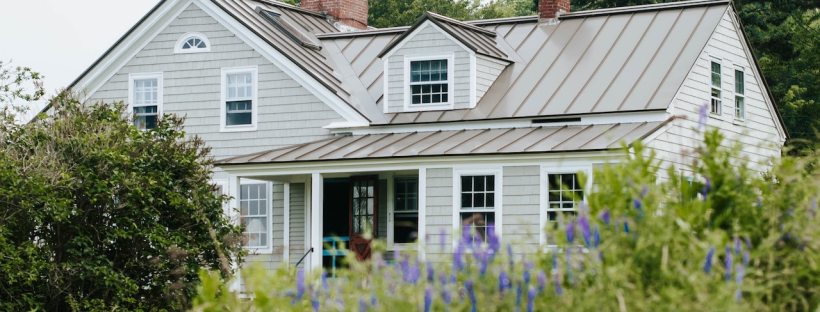 A rather large house with grey siding and a beige roof sits by itself surrounded by greenery and small purple flowers.