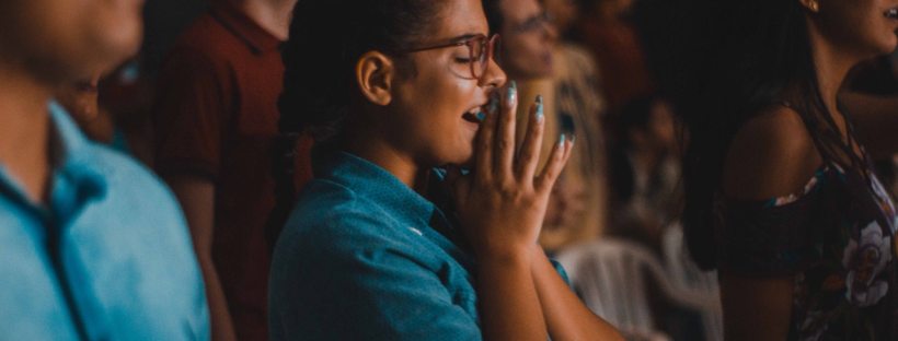 A women stands in a crowd of people singing with her eyes closed and her hands making a prayer sign.