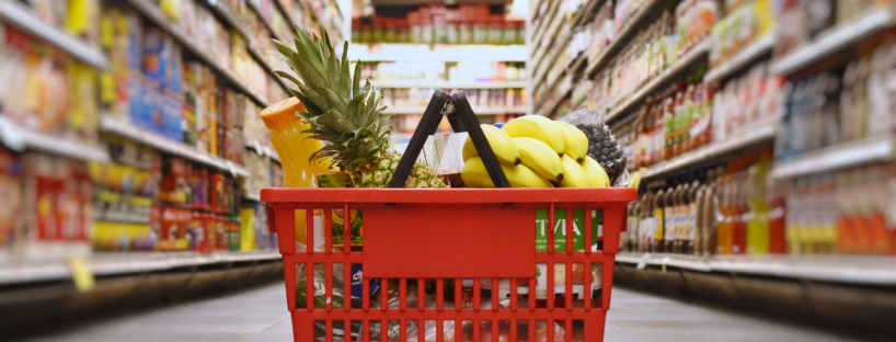 A red basket of groceries—with orange juice, a pineapple, bananas, blueberries and yogurt, among other things—sits in the middle of a grocery store aisle.