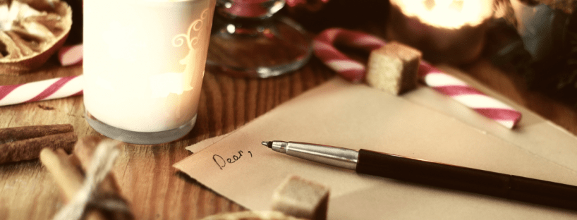 A pen sits on a piece of paper with the word "Dear," started. The table is covered in candles, candy canes, cinnamon and other festive items.