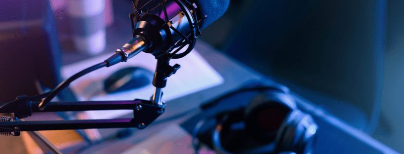 A microphone and a laptop with headphones sit on a desk in a dimly lit room,