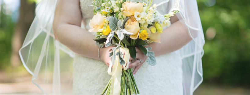 A bride stands in a forest holding her bouquet of yellow, white and green flowers.