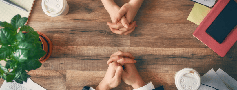 A man and a woman (hands only) sit across from each other at a desk, hands clasped. They each have a coffee, notebooks and charts.