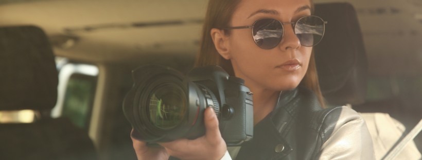 A woman in sunglasses with a camera sits in a car on what looks like a stakeout