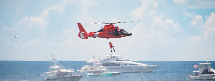 An orange coast guard helicopter is hovering over some big white boats while a member of the Coast Guard is lowered toward them