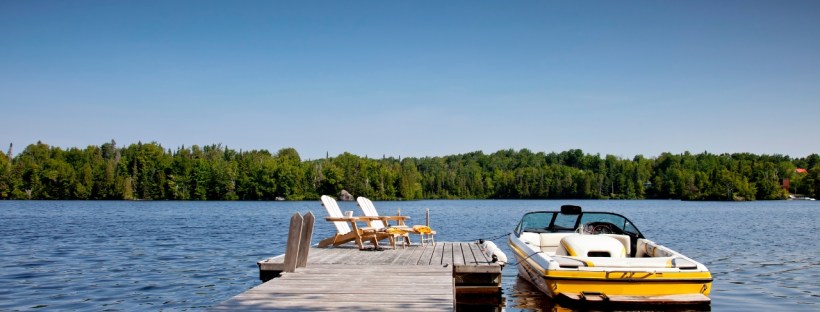 A yellow boat sits on a dock on a lake with Muskoka chairs